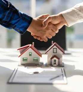 Zwei Personen schütteln sich über einem Schreibtisch die Hände. Auf dem Schreibtisch liegen zwei kleine Modellhäuser und ein Dokument mit einem Stift. Der Händedruck deutet auf eine Vereinbarung oder einen Immobiliendeal hin.